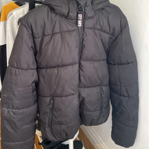 Svart pufferjacka från City Days - Svart pufferjacka från City Days med huva och dragkedja framtill. Jackan har två fickor med dragkedja och quiltad design som ger en cool och modern look. Perfekt för kalla dagar när du vill hålla stilen enkel och snygg.