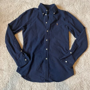 Marinblå skjorta från Ralph Lauren  - Klassisk marinblå skjorta från Ralph Lauren i bomull med button-down krage och broderad logga på bröstet. Custom fit-modell med vita knappar och lång ärm. Perfekt för en stilren och tidlös look.