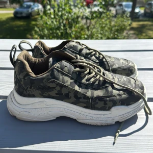 Chunky sneakers med camouflage-mönster - Snygga chunky sneakers med grön och svart camouflage-mönster och vita, tjocka sulor. Skorna har snörning och rund tå, samt dragflik bak för enkel påtagning. Perfekta för dig som gillar streetstyle och vill sticka ut lite extra.