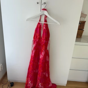 Rosa och röd långklänning Boohoo - Snygg långklänning från Boohoo i rosa och rött med batikmönster. Djup v-ringning, tunna axelband och öppen rygg. Klänningen har snörningsdetaljer i midjan och är tillverkad i ett lätt, flowy material. Perfekt för sommarens alla tillfällen!