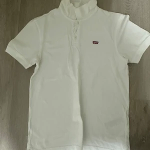 Vit piké t-shirt från Levi's - Vit piké t-shirt från Levi's med klassisk krage och knappar framtill. Liten röd Levi's-logga på bröstet. T-shirten har korta ärmar och är tillverkad i mjuk bomull, perfekt för en clean och stilren look.