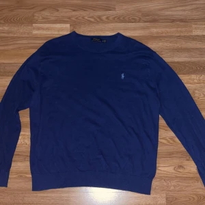 Blå stickad tröja från Polo Ralph Lauren - Mörkblå stickad tröja från Polo Ralph Lauren med klassisk rund halsringning och broderad logga på bröstet. Tröjan har lång ärm och är tillverkad i mjukt material som passar perfekt för lager-på-lager-stil.