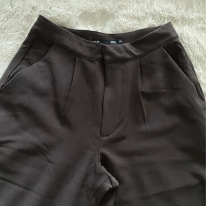 Bruna vida kostymbyxor från 157 XS - Mörkbruna kostymbyxor från 157 i storlek XS. Byxorna har hög midja, vida ben och pressveck framtill. Klassisk design med fickor i sidorna och en bakficka. Perfekta för en clean och stilren look.