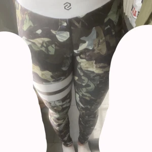 Camo leggings från STRONGER - Snygga leggings från STRONGER med camouflagemönster i grönt, beige och svart. Hög midja med vit linning och vita detaljer på ena benet. Perfekta för träning eller chill. Stretchigt syntetmaterial som sitter tight och bekvämt. Använda en gång och är i väldigt bra skick.