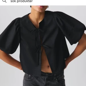 Svart blus med puffärm från Gina Tricot - Svart blus från Gina Tricot med korta puffärmar och två knytningar framtill. Blusen har en rak, croppad passform och är tillverkad i ett slätt, lätt glansigt tyg. Perfekt för dig som gillar trendiga detaljer och stilren look.