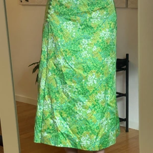 Grön blommig långkjol med knappar - Superfin långkjol i grönt med blommigt mönster och små vita och gula detaljer. Kjolen har en rad klädda knappar längs sidan och är lätt utsvängd för en flowy känsla. Perfekt för dig som vill sticka ut med färg och mönster i sommar.