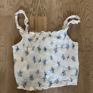 Blommig smockad linnetopp  - Supersöt vit linnetopp  med blått blommönster och smockad struktur. Toppen har smala axelband med rynkade detaljer och vågig kant upptill och nedtill. Perfekt för varma dagar och ger en fräsch sommarkänsla.