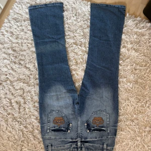Blå bootcut jeans med broderi - Snygga blå bootcut jeans från Chang Ying Jeans med broderade detaljer på bakfickorna och ett coolt ansiktsmotiv med nitar på ena låret. Jeansen har klassisk femficksdesign, knapp och dragkedja framtill samt slitningar vid bensluten. Kan sälja billig skriv bara för förslag på pris! 💕