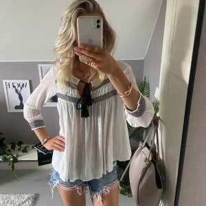 Vit blus med svarta detaljer från Bershka - Luftig vit blus från Bershka med svarta broderade detaljer och knytband med tofsar vid halsen. Blusen har trekvartsärm och är tillverkad i ett tunt, skönt material som ger en avslappnad vibe. Perfekt till jeansshorts eller kjol för en chill look.