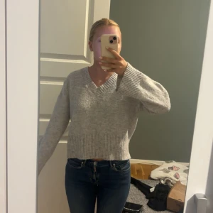 Grå stickad croppad tröja med v-ringning - Mysig grå stickad tröja med v-ringad hals och croppad passform. Tröjan har breda ribbade muddar vid ärmslut och nederkant, samt långa ärmar. Perfekt för lager-på-lager under kyliga dagar.