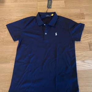 Marinblå pikétröja från Ralph Lauren - Klassisk marinblå pikétröja från Ralph Lauren med vit broderad logga på bröstet. Tröjan har korta ärmar, krage och knäppning med tre knappar. Perfekt för en stilren och sportig look.