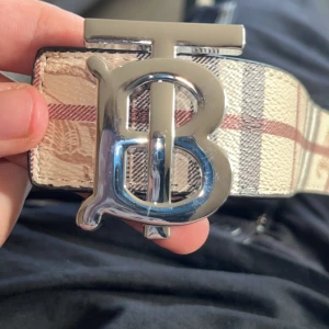 Burberry bälte med rutigt mönster - Snyggt Burberry bälte med klassiskt rutigt mönster i beige, svart, vitt och rött. Bältet har en stor silvrig spänne med TB-logga och är gjort i skinn. Perfekt accessoar för att lyfta din outfit med en lyxig touch.