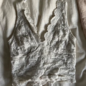 Vit spetsig linnetopp från Amisu - Supersöt vit linnetopp från Amisu i storlek XS. Toppen är täckt av vacker spets med blommigt mönster och har djup v-ringning samt ärmlös design. 