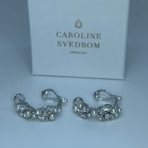 Caroline Svedbom silvriga örhängen - Snygga silvriga örhängen från Caroline Svedbom med oregelbunden form, dekorerade med små vita pärlor och glittrande stenar. Fått dom i studentpresent men säljer dom då jag inte är en silvertjej, priset är 500kr🫶🏽