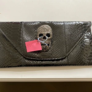 Svart kuvertväska med döskalle - Cool svart kuvertväska i glansigt skinnimitation med ormmönster och en blingad döskalle på locket. Perfekt för dig som vill sticka ut och gillar edgy detaljer. Väskan har ett stilrent kuvertformat och stängs med lock.