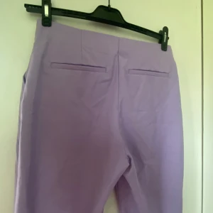 Ljuslila bootcut kostymbyxor - Snygga ljuslila kostymbyxor med bootcut-ben och hög midja. Byxorna har två fusk-bakfickor och sidfickor, perfekta för en trendig och färgglad look. Materialet känns som bomull eller polyester, vilket ger en mjuk och bekväm känsla.