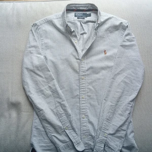  skjorta  Polo Ralph Lauren  - Snygg smalrandig skjorta från Polo Ralph Lauren i vitt och blått. Slim fit-modell med klassisk krage, långa ärmar och knäppning framtill. Broderad logga på bröstet. Perfekt för dig som gillar stilrena och tidlösa plagg.