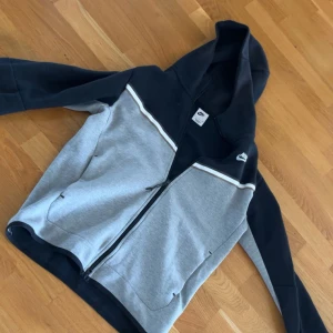 Nike hoodie i grått och svart - Nike hoodie med dragkedja i grått och svart. Tröjan har huva, långa ärmar och en vit rand över bröstet. Perfekt för chill dagar eller när du vill ha en sportig look. Tillverkad i mjuk bomullsblandning för extra komfort.