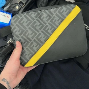 Fendi axelväska med gul detalj - Snygg axelväska från Fendi med klassiskt FF-mönster i grått och svart, samt en diagonal gul skinnpanel. Väskan har en stilren, rektangulär form och justerbar axelrem i svart. Perfekt för dig som vill sticka ut med en ikonisk designeraccessoar.