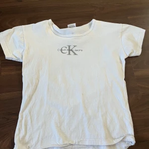 Vit t-shirt från Calvin Klein Jeans - Vit t-shirt från Calvin Klein Jeans med klassisk CK-logga tryckt på bröstet. Enkel och stilren design med rund halsringning och korta ärmar. Tillverkad i mjuk bomull som känns skön mot huden. Perfekt för en clean och avslappnad look.