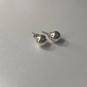  silverfärgade pärlörhängen - Säljer ett par stilrena örhängen i form av blanka, runda bollar i silverfärg. De har klassisk stiftmodell och passar perfekt om du gillar minimalistiska smycken med en modern touch. Enkla att matcha med allt och ger en clean look.