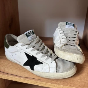 Golden Goose🪖 - Golden Goose sneakers i vitt skinn med grå mockadetaljer, svart stjärna på sidan och mörkgröna hälkappor. Klassisk låg modell med snörning och platt sula. Dojjorna är i väldigt bra skick, behöver endast en lätt tvätt. Jag är villig att gå ned lite i pris vid snabb affär! Samt tar även emot bytesförslag!🙌🏼 OBS! Lådan finns tyvär inte med.