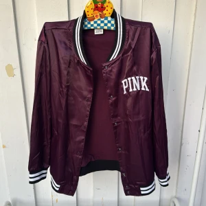 Vinröd bomberjacka från PINK - Snygg vinröd bomberjacka från PINK med vita detaljer och svartvita ribbade muddar. Jackan har tryckknappar framtill, broderad PINK-logga på bröstet och klassisk bomberkrage. Perfekt för dig som vill ha en sportig och trendig look.