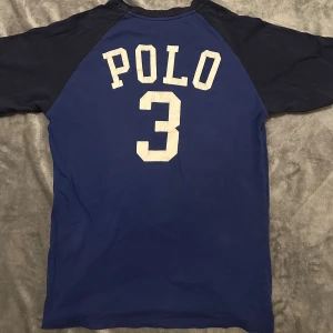 Blå t-shirt Polo Ralph Lauren - Mörkblå t-shirt från Polo Ralph Lauren med kontrasterande svarta ärmar och vit logga på bröstet. Baksidan har stort tryck med texten 'POLO' och siffran 3. Klassisk rund hals och sportig vibe. Tillverkad i mjuk bomull.