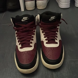 Nike sneakers i vinrött och vitt - Snygga Nike sneakers med högt skaft i vinrött, vitt och svart. Klassisk design med svart swoosh, perforerad tå och vinröda skosnören. Ovandel i syntet och skinn, perfekt för dig som gillar streetstyle och vill sticka ut med färg.