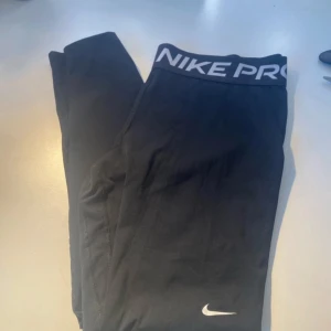 Nike Pro svarta tights  - Svarta Nike Pro tights med bred resår i midjan och vit logga på benet. Tillverkade i stretchigt Dri-FIT-material som andas. Perfekta för träning och sportiga aktiviteter. Sitter tight och följer kroppen för maximal rörelsefrihet.
