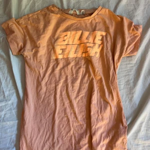Billie Eilish  t-shirt - Peachfärgad t-shirt med tryck av Billie Eilish-loggan i orange på bröstet. Klassisk rund hals och korta ärmar. Jätte fin och skön att ha på sig.