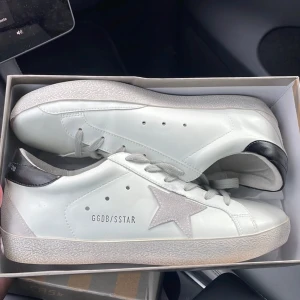 Vita sneakers från Golden Goose med silverstjärna - Säljer ett par vita sneakers från Golden Goose med silverfärgad stjärndetalj på sidan och svart hälparti. Skorna har snörning och platt sula, samt GGDB/SSTAR-tryck på sidan. Perfekta för en stilren och trendig look. Helt fabriksnya och aldrig använda 