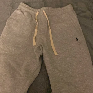 Grå mjukisbyxor från Polo Ralph Lauren - Snygga ljusgrå mjukisbyxor från Polo Ralph Lauren med dragsko i midjan och broderad logga på ena benet. Byxorna har fickor på sidorna och en bakficka. Perfekta för en avslappnad stil.