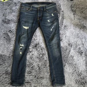Blå slitna jeans från Nudie Jeans - Säljer ett par blå jeans från Nudie Jeans med slitna detaljer och lagningar på jeansen, nyskick använd ett fåtal gånger, nypris-1800 mitt pris 750 fler bilder kan självklart fås.