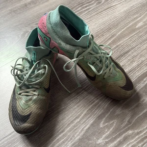 Nike Mercurial fotbollsskor i mintgrön och rosa - Säljer ett par Nike Mercurial fotbollsskor i mintgrön med svarta detaljer och rosa inslag på hälen. Skorna har snörning, hög krage och är tillverkade i syntetmaterial. Perfekta för dig som vill sticka ut på planen!