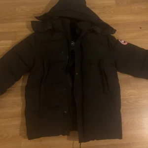 Canada goose - Säljer en svart dunjacka från Canada Goose med huva och klassisk logotyp på ärmen. Jackan har lång ärm, dragkedja och knappar framtill samt två stora fickor. Perfekt för kalla dagar.
