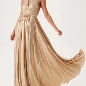 Guldig långklänning med djup v-ringning - Säljer en elegant guldig långklänning med djup v-ringning och öppen rygg. Klänningen har ett plisserat, glansigt tyg som ger ett lyxigt intryck och ett brett midjeband som markerar midjan. Perfekt för bal eller festliga tillfällen. Aldrig använd och prislapp sitter kvar