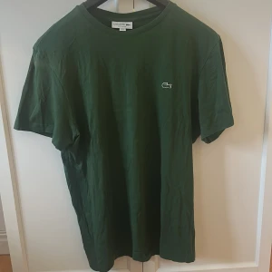 Grön t-shirt från Lacoste - Säljer en klassisk grön t-shirt från Lacoste med rund hals och liten broderad logga på bröstet. T-shirten har normal passform och är tillverkad i mjuk bomull. Perfekt för en stilren och enkel look.