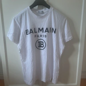 Vit t-shirt från Balmain  - Säljer en vit t-shirt från Balmain med svart tryck framtill. Klassisk rund halsringning och korta ärmar. Perfekt för dig som gillar stilrena och exklusiva plagg med tydlig logga.