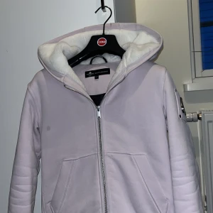 Ljuslila hoodiejacka från Moose Knuckles - Säljer en ljuslila hoodiejacka från Moose Knuckles med dragkedja och två fickor framtill. Jackan har en mysig vit fleecefodrad huva och ikonisk logga i metall på ärmen. Perfekt för kyliga dagar.