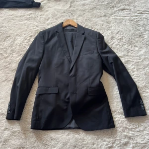 Svart kavaj från H&M - Klassisk svart kavaj från H&M i regular fit. Kavajen har två framfickor med lock, bröstficka och knappar vid ärmslut. Perfekt för bal eller fest. Tillverkad i polyesterblandning med slät finish.