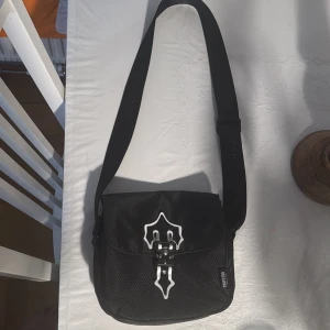 Trapstar Väska - Säljer en svart axelväska från Trapstar med justerbar rem och unikt spänne framtill i svart och vitt. Väskan har ett coolt mönster och logga på framsidan. Perfekt för att bära dina viktigaste grejer med stil.