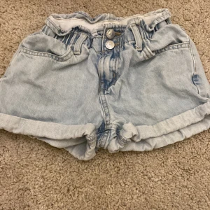 Ljusblå jeansshorts från Lindex  - Ljusblå jeansshorts från Lindex med midja och dubbla knappar framtill. Shortsen har uppvikta benslut och två bakfickor. Perfekta för varma dagar och enkel att matcha med olika toppar.
