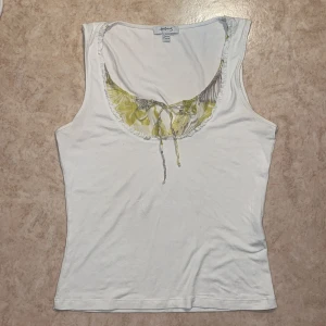 Vit ärmlös topp med blommigt mönster och knytning - Säljer en vit ärmlös topp med blommigt mönster i gult och grönt vid urringningen. Toppen har en söt knytning framtill och är i mjuk bomull. Köpt secondhand i Spanien, har en nästintill osynlig fläck på baksidan, men annars i bra skick! 
