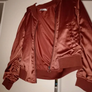 Satin bomberjacka från Gina Tricot - Säljer en vinröd/korall färgad bomberjacka i glansigt satinmaterial från Gina Tricot. Jackan har dragkedja framtill, ribbade muddar och rynkade detaljer längs ärmar och rygg. Perfekt för dig som vill sticka ut med en unik jacka! ‼️Liten i storlek mer som M/L än XXL‼️