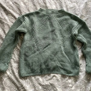 Grön stickad tröja från Hollys - Mysig grön stickad tröja från Hollys i storlek M. Tröjan har ett grovt stickat mönster och långa ärmar med ribbade muddar vid hals, ärmslut och nederkant. Perfekt för kyligare dagar.