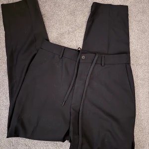 These Glory Days Svarta kostymbyxor med snörning - Säljer ett par svarta kostymbyxor med snörning i midjan och knappstängning. Byxorna har raka ben. . Passar dig som gillar klassiska och enkla plagg.