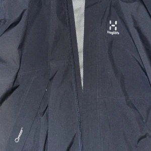 Mörkblått skaljacka från Haglöfs - Svart skaljacka från Haglöfs med diskret logga på bröstet och dragkedja framtill. Jackan är tillverkad i vattentätt material och har minimalistisk design, perfekt för utomhusaktiviteter. XL och är lite lång men kan också  justera pass formen. Har fått den som en present och just att den är lite för stor för mig därför säljer jag den. Ingen Frakt❌