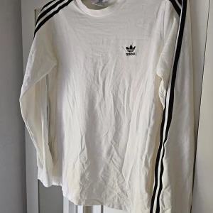 Vit långärmad tröja från Adidas stl M  - Säljer en klassisk vit långärmad tröja från Adidas med svarta ränder längs ärmarna och liten Adidas-logga på bröstet. Tröjan har rund hals och är tillverkad i mjuk bomull. Perfekt för en sportig och avslappnad stil. Storlek M