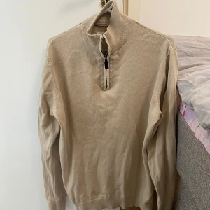 Beige stickad tröja med dragkedja - Säljer en beige stickad tröja med halv dragkedja i halsen. Tröjan har lång ärm och ribbade muddar vid ärmslut och nederkant. Perfekt för lager-på-lager och enkel att matcha med olika outfits. Storlek L  känns som M